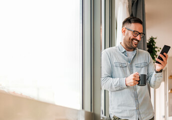 Naklejka na ściany i meble Smiling man businessman in casuals standing in office next to window using mobile phone drinking coffee holding coffee cup Small business entrepreneur looking at smart phone while taking coffee break