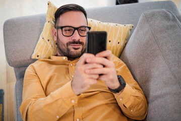 Cheerful young adult mature man happy smiling using smart phone resting lying down on sofa couch at home reading messages on mobile phone Guy with eyeglasses relaxing at home surfing net social media © Moon Safari