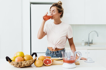 Woman drinking freshly squeezed homemade grapefruit juice in white kitchen - obrazy, fototapety, plakaty