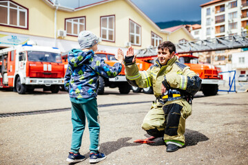 Obraz premium Portrait of a firefighter with kid standing against the backdrop of fire trucks. Childs dreamed profession. Fire station