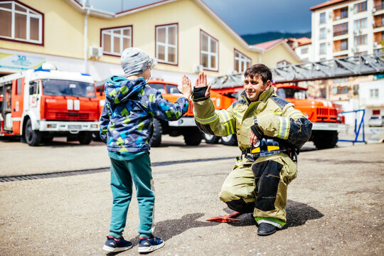 Portrait of a firefighter with kid standing against the backdrop of fire trucks. Childs dreamed profession. Fire station