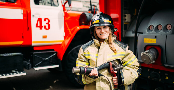 Smiling female firefighter in helmet looking at camera behind at fire engine. Fire station
