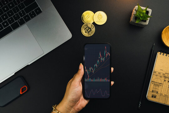 Girl holding a smartphone with financial stock market graph on the screen on black table with a cryptocurrencies coins and computer. Office environment. Stock Exchange