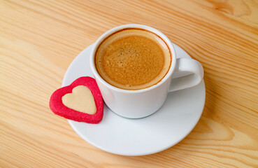A cup of hot coffee with a heart shaped cookie on wooden background
