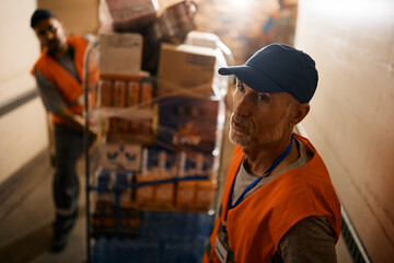 Mature warehouse worker and his coworker moving rack with packages through storage hallway.