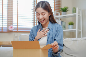 Happy excited, asian young woman, girl customer sitting on sofa at home, opening and unpacking cardboard box carton parcel after buying ordering present, shopping online, delivery service concept.