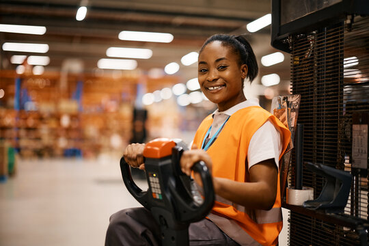 Happy African American woman driving pallet jack while working at distribution warehouse.