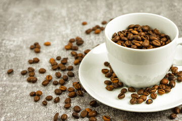 White cup of coffee with coffee beans on gray concrete background with scattering of coffee beans. Arabica, Robusta. Drink for office and home. Background for site with copy space for text