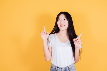 Smiling Asian woman with long dark hair wears white t-shirt pointing finger to the side on yellow background.