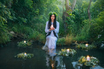fantasy pagan witch slavic woman holding candle in hand. Divination summer tradition. Girl Mermaid nymph sits on banks river. White vintage nightgown dress. summer nature green grass trees. Long hair - obrazy, fototapety, plakaty