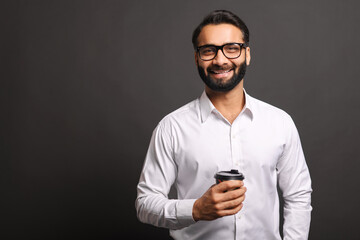 Handsome confident bearded Indian man in formal wear stands with paper cup of coffee to-go isolated on black, smiling male office employee in glasses take a coffee break
