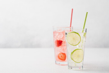 Lime mojito and strawberry lemonade with ice in tall glasses on light gray background. Berry refreshing summer drinks with straw