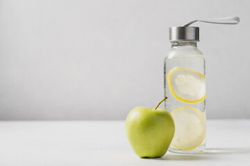 Fitness or diet concept. Refreshing water with lemon in glass bottle and green apple on light gray background with copy space