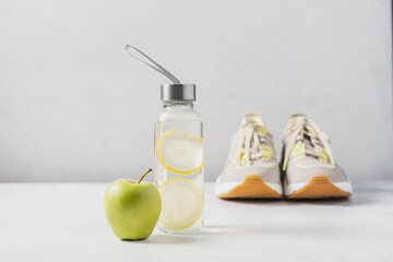 Fitness concept. Refreshing water with lemon in glass bottle and green apple with sports sneakers in the background