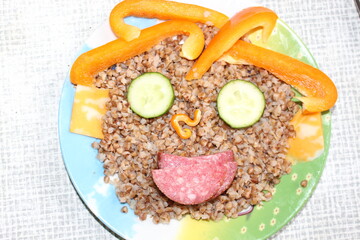 Breakfast for a child is decorated in the form of a funny face in a plate. Buckwheat, pepper, cucumber, cheese, sausage are good for the body