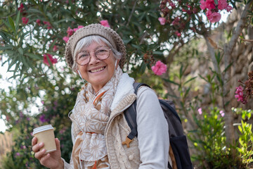 Portrait of senior attractive woman wearing glasses and cap sitting outdoors in public park holding a coffee cup while listening to music by earphones