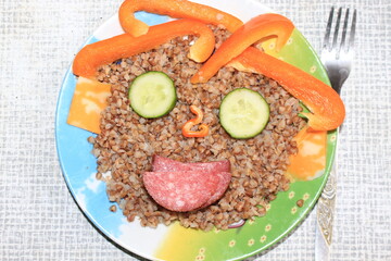 Breakfast for a child is decorated in the form of a funny face in a plate. Buckwheat, pepper, cucumber, cheese, sausage are good for the body