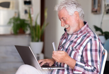 Elderly bearded man browsing by laptop enjoying shopping online. Joyful and smiling beautiful senior man at home being in great mood using credit card to purchase