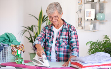 Smiling elderly woman in checkered shirt and glasses ironing clothes at home on ironing board