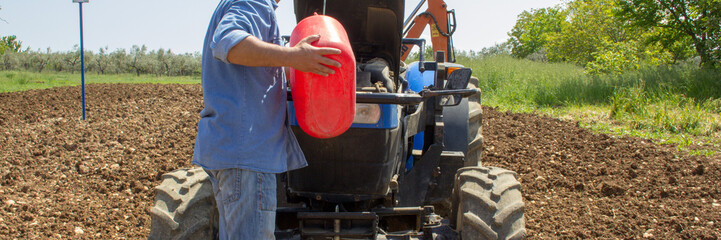 Farmer refueling his tractor with a jerry can, as he runs out of fuel. Reference to the expensive...