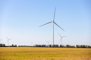 Panoramic landscape view of new white modern wind turbine farm power generation station against clear blue sky and field. Clean sustainable zero emission alternative electricity windfarm industry