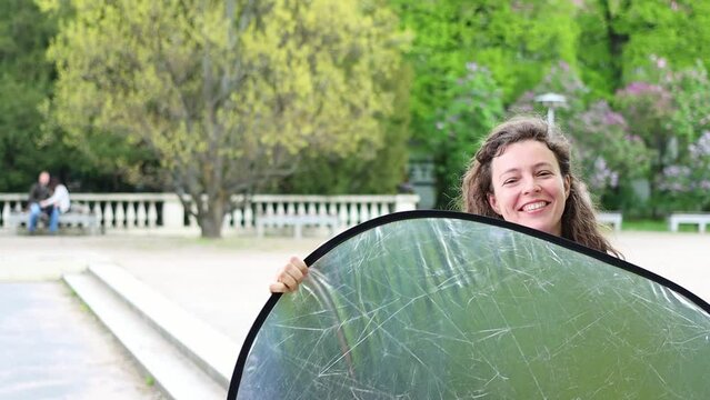 Happy photographers assistant holding reflector helping to control light over model at location shoot outdoors in city park location.Smiling young girl student taking a photography course.Slow motion