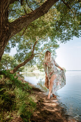 Fototapeta premium A young 30 year old woman in a long dress and barefoot walks along the beach lake. Beautiful landscape. Feminine style of a European girl in sunglasses and straw hat. Slim body. Natural environment