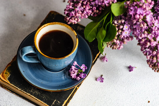 Cup of coffee on a hardback book next to a vase with lilac flowers