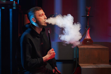 a young man in profile smokes a hookah, exhaling a lot of smoke in a dark room