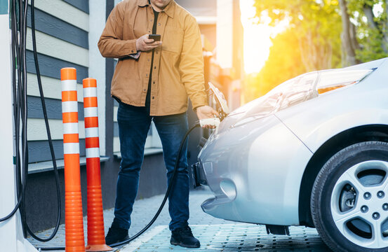 Young man traveling by electric car at summer, having stop at charging station standing plugging cable and browsing internet on smartphone joyful while charing