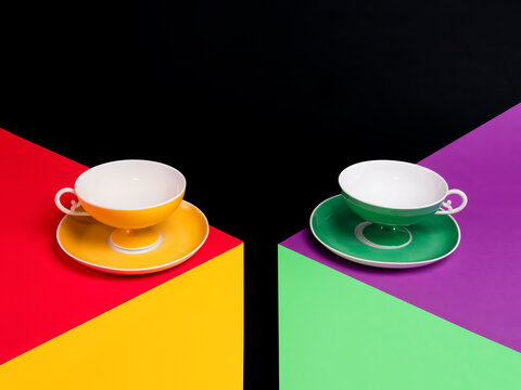 Two colourful empty teacups levitating off saucers on two multi coloured tables