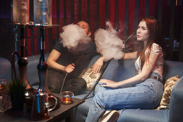 a young woman and a young man smoke a hookah while sitting on a sofa and exhaling thick white...