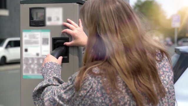 Eine Frau steht an einer E-Auto Ladestation. Sie steckt das Ladekabel vom Typ 2 in eine &ouml;ffentliche Ladestation. Im Hintergrund ist eine Stra&szlig;e mit Verkehr. Echtzeit.