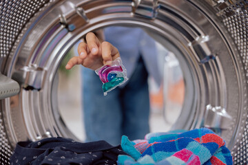 A woman puts a capsule of liquid powder into a washing machine with laundry, close-up view from...
