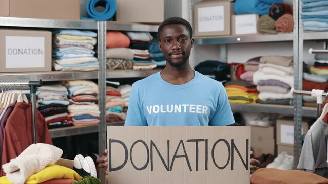 Portrait of energetic multiracial man looking at the camera and smiling while holding banner with donation word in charity shop. Volunteers working in organization and sorting donated clothes