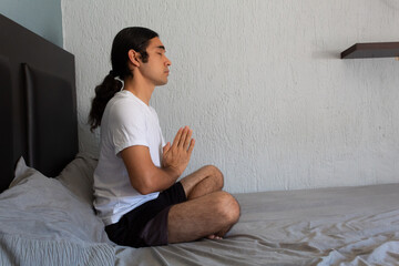 Hispanic non binary long-haired man doing yoga and meditating on his bed