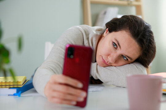 Bored freelancer woman sitting at the table at home, using smartphone when working
