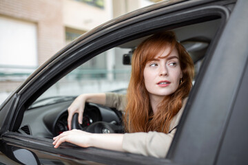 Naklejka na ściany i meble Joyful redhead woman inside of car looking back from driver seat while driving during the day.