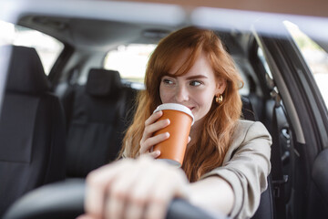 Happy young woman with coffee to go driving her car. Woman sipping a coffee while driving a car. Young woman drinking coffee while driving her car. Attractive red hair drives a car - Powered by Adobe