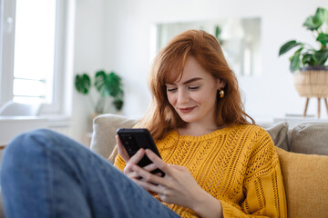 Smiling pretty young woman, relaxes on the sofa in her living room while using her mobile smart phone for social media and surfing the internet