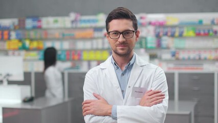 Handsome caucasian man in eyeglasses and white lab coat smiling and looking at camera while posing at drugstore with crossed arms. Female colleague working behind on modern computer.