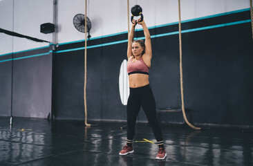 Muscular female bodybuilder in tracksuit training with kettlebell during hard workout in gym studio, strong Caucasian athlete using sportive barbell equipment for exercising during crossfit practice