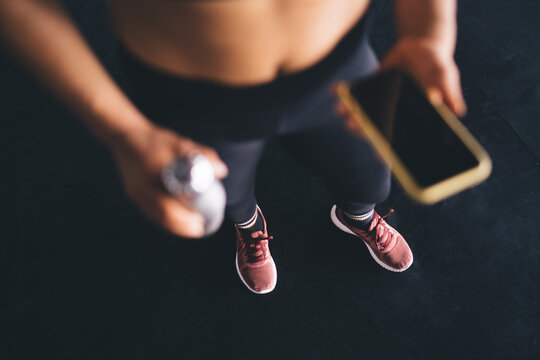 Unrecognizable female athlete in sportive sneakers using blurred cellphone technology for mobile chatting during break for refreshing on workout training, cropped woman with smartphone in gym studio