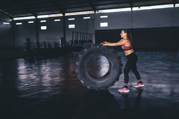 Fototapeta na wymiar Cheerful woman 20 years old working out with training equipment during time in sport hall doing cardio exercises, smiling female athlete with wheel tire weightlifting in gym studio interior