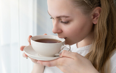 Young woman with a mug of tea in her hands sits near the window, space for text