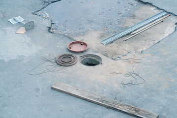 An open round hatch in the ground and metal hatch covers lying with construction junk around. Abandoned territory. Underground engineering works. Repair and construction of underground sewers.