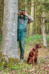 A hunter with dungarees and rubber boots stands behind a tree at the edge of the forest in the late afternoon and watches the game through his binoculars. His Irish Setter hound sits next to him.