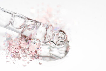 Piipette with transparent liquid on light glass background with splash of shiny pink glitter around and copy space. Texture of face serum or cosmetic oil. Selective focus, macro photo
