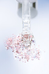 Vertical image of piipette with transparent liquid on light glass background with splash of shiny pink glitter around and copy space. Texture of face serum or cosmetic oil. Selective focus