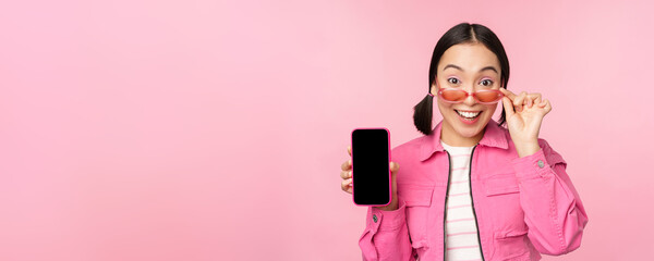 Portrait of asian girl showing mobile phone screen, reacting surprised, standing over pink...